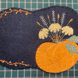 Puede incluir: Un mantel individual de fieltro gris oscuro con una calabaza naranja bordada y un arreglo floral. La calabaza está centrada, con un ramo de flores amarillas y hojas verdes. Un borde de luces naranjas y amarillas decora el lado izquierdo.