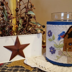 May include: A white planter with a red star on the side, filled with brown branches and green leaves. Next to it is a glass jar with a white felt cover featuring a brown birdhouse with two blue birds and purple flowers.