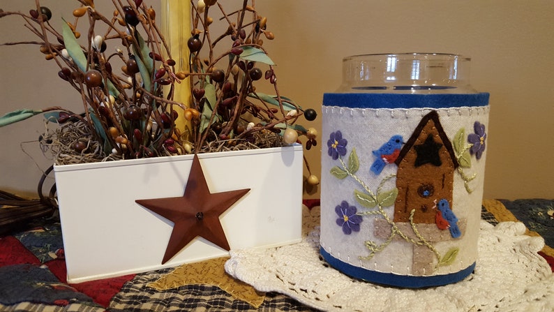 May include: A white planter with a red star on the side, filled with brown branches and green leaves. Next to it is a glass jar with a white felt cover featuring a brown birdhouse with two blue birds and purple flowers.