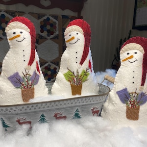 Puede incluir: Tres decoraciones de muñecos de nieve con sombreros y bufandas rojas, cada uno con una cara sonriente y una pequeña cesta de vegetación. Se encuentran en una bandeja de metal con diseños festivos, rodeados de nieve artificial. Un encantador tema de vacaciones de invierno.