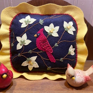 Könnte beinhalten: Ein handgefertigtes Filzkissen mit einem roten Kardinal und weißen Blumen, die auf einem dunkelblauen Hintergrund gestickt sind. Das Kissen hat einen gelben, gewellten Rand. Zwei kleine rote Kardinalfiguren befinden sich im Vordergrund.