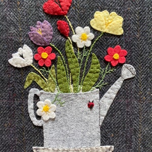 May include: A white watering can with a red ladybug on the side, filled with a bouquet of felt flowers in shades of red, white, yellow, purple, and green. The flowers are arranged in a vintage style.