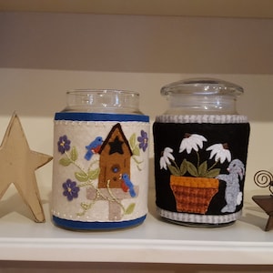 May include: Two glass jars with felt covers. The jar on the left has a blue, white, and brown felt cover with a birdhouse and birds. The jar on the right has a black, white, and brown felt cover with a flower pot and a rabbit.