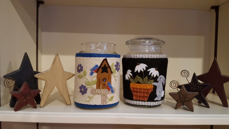 May include: Two glass jars with felt covers. The jar on the left has a blue, white, and brown felt cover with a birdhouse and birds. The jar on the right has a black, white, and brown felt cover with a flower pot and a rabbit.