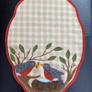 May include: A brown and tan oval-shaped fabric placemat with a red border. Two embroidered birds are perched on a branch above a nest with blue eggs. The birds are gray with red breasts and black eyes.
