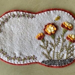 May include: A white felt oval with a red border, featuring embroidered flowers in shades of orange, yellow, and brown. A small bee is also embroidered on the right side of the oval.