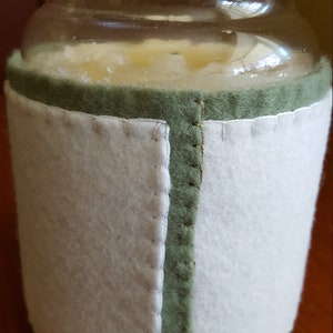 May include: A clear glass candle jar with a white candle inside. The jar is wrapped in a green and white felt cover.