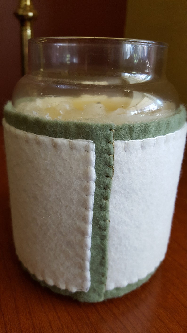 May include: A clear glass candle jar with a white candle inside. The jar is wrapped in a green and white felt cover.