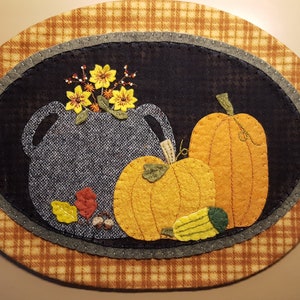 May include: A handmade felt wall hanging with a blue and gray crock, two orange pumpkins, and yellow flowers. The background is a brown and orange plaid.
