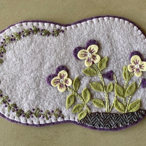 May include: A purple felt oval with a stitched border and a design of three purple and yellow flowers with green leaves. The flowers are embroidered on the felt.