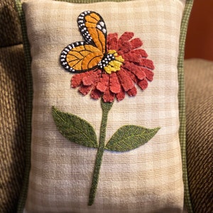 May include: A beige pillow with a green border features an embroidered design of a butterfly landing on a red flower with green leaves and a stem.