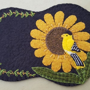 Pode incluir: Uma tapeçaria de parede de feltro com um girassol amarelo e um pequeno pássaro amarelo com listras pretas. O girassol é cercado por folhas verdes e uma coroa de folhas verdes com bagas laranja.