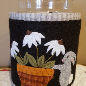 May include: A black felt jar cozy with a white bunny rabbit peeking out from behind a potted plant with white flowers. The pot is brown with a checkered pattern. The cozy has a white and gray checkered trim.