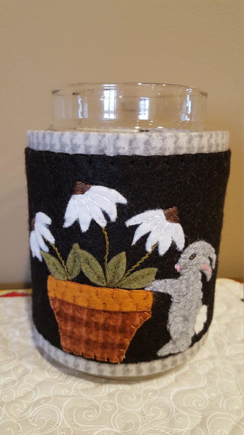 May include: A black felt jar cozy with a white bunny rabbit peeking out from behind a potted plant with white flowers. The pot is brown with a checkered pattern. The cozy has a white and gray checkered trim.