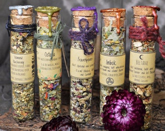 Può includere: Cinque piccoli flaconi di vetro con tappi di sughero e sigilli di cera. Ogni flacone contiene erbe e fiori secchi. Le etichette sui flaconi sono in tedesco e elencano i nomi delle erbe e i loro usi. I flaconi sono disposti in fila su una superficie di legno.
