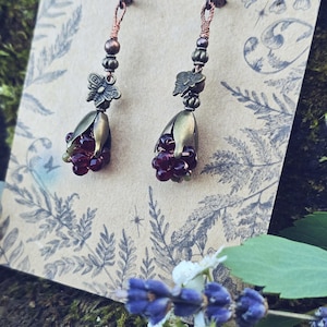 Può includere: Un paio di orecchini fatti a mano a tema botanico. Ogni orecchino presenta un gruppo di piccole perline rosso scuro all'interno di un bocciolo di fiore color ottone. Gli orecchini sono adornati da un ciondolo a farfalla e sono appesi a un filo di rame.
