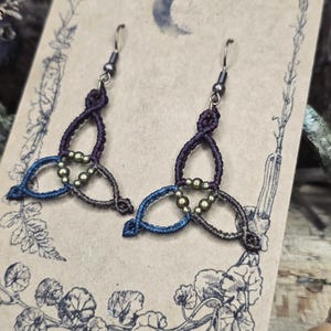 Puede incluir: Un par de pendientes de macramé hechos a mano con un diseño de nudo celta. Los pendientes están hechos con cordón morado oscuro, azul y marrón, acentuados con pequeñas cuentas doradas. Cuelgan de ganchos plateados.