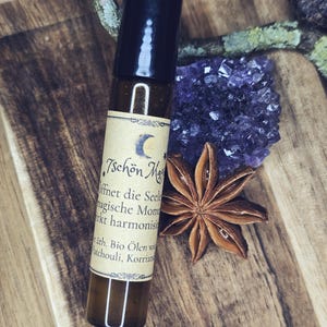 Könnte beinhalten: Eine braune Glas-Ätherisch-Öl-Rollerflasche mit schwarzem Deckel und einem Etikett mit deutschem Text. Die Flasche befindet sich neben einem lila Amethystkristall und einer Sternanis-Schote auf einer Holzoberfläche.