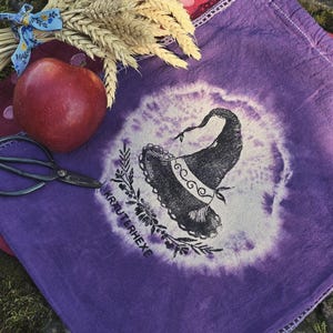 Könnte beinhalten: Ein violetter Stoffbeutel mit Kordelzug, der eine schwarze Druckgrafik eines spitzen Hutes, florale Elemente und den Text KRAUTERHEXE zeigt. Der Beutel hat ein kreisförmiges Batikmuster in der Mitte und einen dekorativen Häkelrand am oberen Saum.