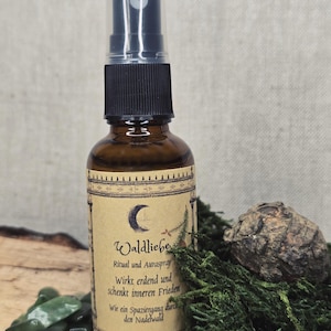 Puede incluir: Un frasco rociador de vidrio marrón con una tapa negra y una etiqueta con texto y un diseño de media luna. La botella está junto a piedras verdes, musgo y una piña. La etiqueta dice "Waldliebe".