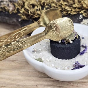 Könnte beinhalten: Nahaufnahme von zwei verzierten, goldfarbenen Zangen mit floralen Designs, die auf einer kleinen weißen Schale liegen. Die Schale enthält Sand, getrocknete Blumen und eine schwarze Holzkohlescheibe. Der Hintergrund zeigt getrocknete Pflanzen auf einer Holzoberfläche.