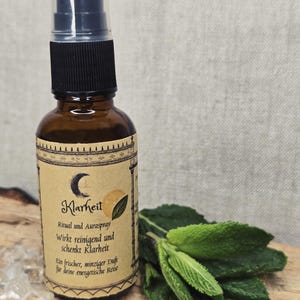 Könnte beinhalten: Eine braune Glassprühflasche mit schwarzem Verschluss und Etikett mit Text und Monddesign. Frische grüne Blätter und klare Kristalle sind neben der Flasche angeordnet. Das Etikett lautet "Klarheit".