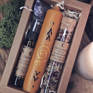 Può includere: Una confezione regalo contenente una candela arancione con dettagli dorati e simboli neri, un piccolo flacone spray, una fiala di vetro con un tappo di sughero e un cristallo viola. La candela ha il testo "Seelen" e "irkt stress und entspa". La fiala ha il testo "Seelenwohl".