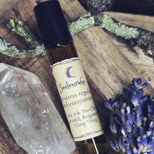 Könnte beinhalten: Eine braune Glas-Rollerflasche für ätherische Öle mit schwarzem Deckel, beschriftet mit Text und einem Mond. Die Flasche ist von Lavendel, einem klaren Kristall und Holzelementen umgeben. Das Etikett lautet "Seelenruhe".