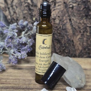 Könnte beinhalten: Eine kleine braune Glas-Ätherisch-Öl-Rollerflasche mit schwarzem Verschluss und einem Etikett mit Text. Die Flasche befindet sich neben einem schwarzen Verschluss, klaren Kristallen und einem Zweig lila Blumen. Das Etikett trägt das Wort "Eiswind".