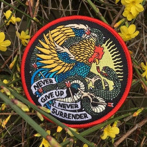 Può includere: Una toppa rotonda ricamata con un bordo rosso. La toppa presenta un gallo che combatte un serpente con il testo "Never Give Up Never Surrender" in uno stendardo attorno all'immagine.
