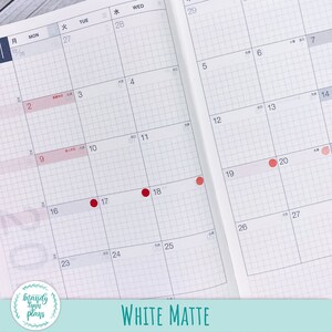 May include: A white matte planner with a grid pattern and red dots marking dates. The planner has Japanese text for the days of the week and numbers for the dates. The planner is open to the month of January.
