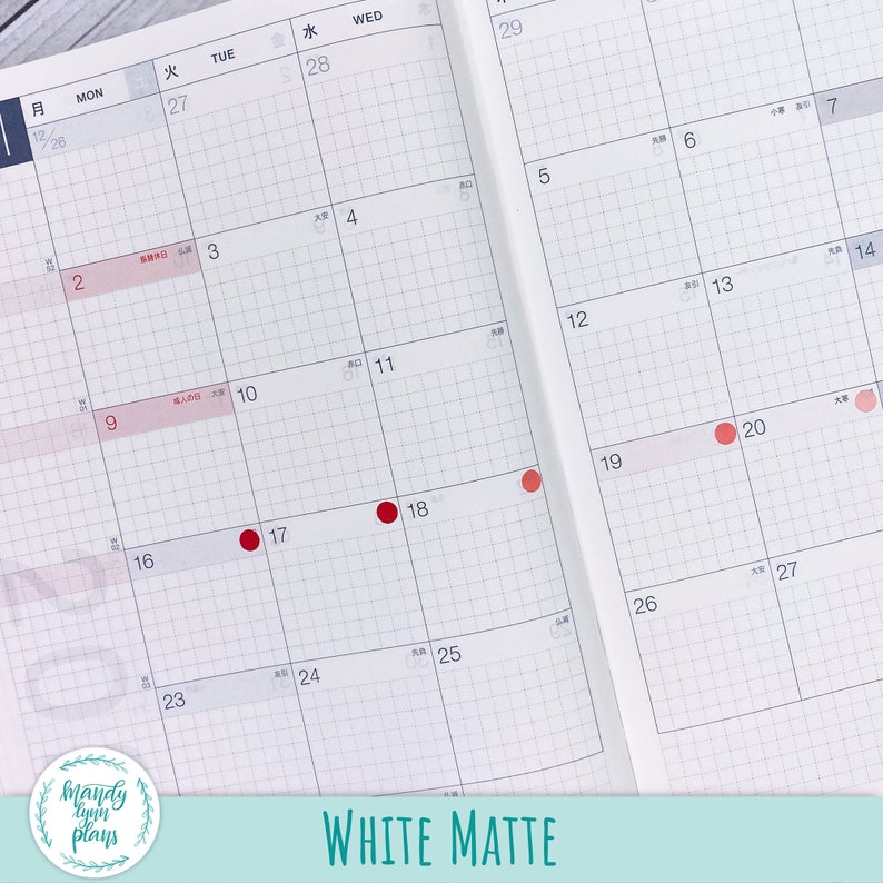 May include: A white matte planner with a grid pattern and red dots marking dates. The planner has Japanese text for the days of the week and numbers for the dates. The planner is open to the month of January.