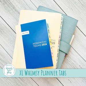 May include: Blue planner with the text "HOBONICHI TECHO 2024" and "day-free" on the cover. The planner has colourful tabs with month names written on them. The text "XL WHIMSY PLANNER TABS" is written on a blue banner.