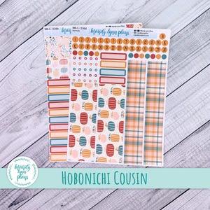 May include: A set of planner stickers with a fall theme. The stickers feature pumpkin and plaid patterns in orange, teal, and pink. There are also date stickers and label stickers. The text "HOBONICHI COUSIN" is at the bottom.