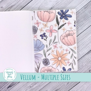May include: Open notebook with grid paper on the left and a floral vellum cover on the right. The vellum features a pattern of pink, blue, and peach flowers with green leaves. Text on the bottom reads "VELLUM - MULTIPLE SIZES".