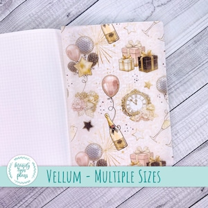 May include: A white notebook with a gold and pink patterned vellum cover. The cover features a repeating pattern of champagne bottles, clocks, stars, balloons, and gift boxes. The text "VELLUM - MULTIPLE SIZES" is printed on the bottom of the image.