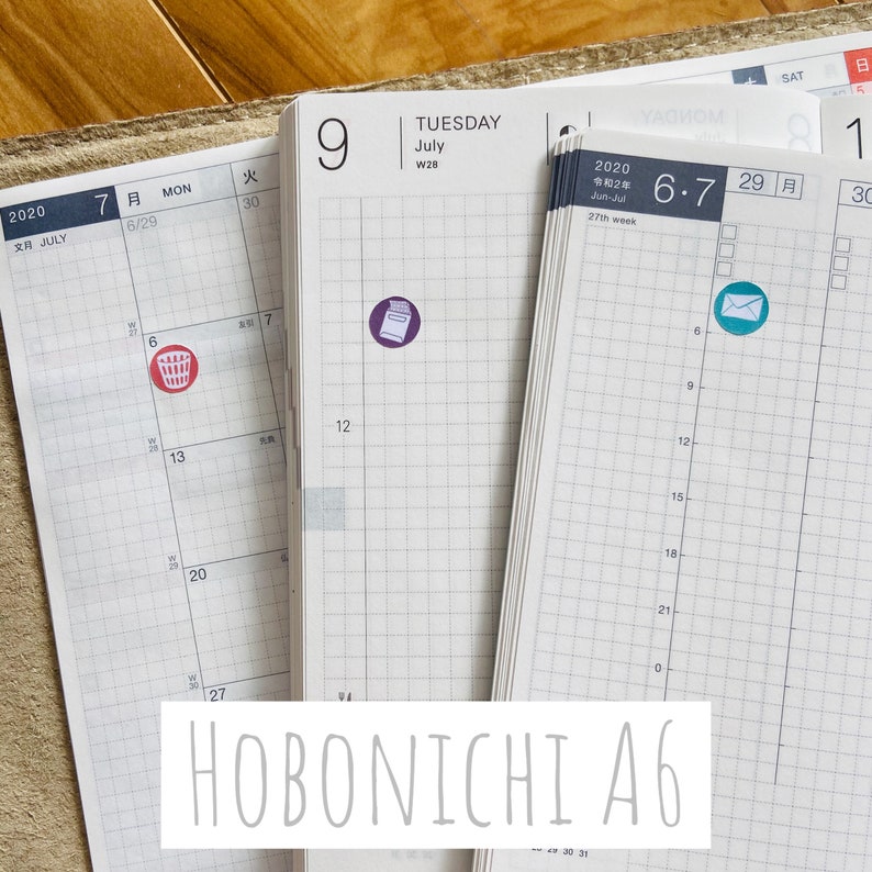 May include: A close-up of a Hobonichi A6 planner with two stickers. The left sticker is red and depicts a trash can. The right sticker is blue and depicts an envelope. The planner is open to July 2020.