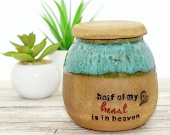 May include: A handcrafted ceramic urn with a teal and brown glaze. The urn has a lid and features the text "half of my heart is in heaven" in black and red. A decorative item for remembrance.