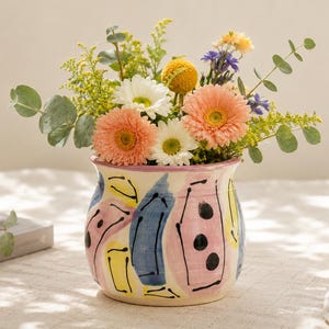 May include: A colorful ceramic vase with a hand-painted design, filled with a vibrant arrangement of fresh flowers. The vase features pink, blue, and yellow abstract shapes and black dots. The flowers include orange gerbera daisies, white daisies, and yellow and purple accents.