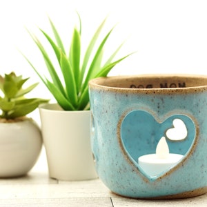 May include: Teal blue ceramic candle holder with a heart-shaped cutout and the words "Dog Mom" on the rim. The holder is sitting on a white surface with two potted plants.
