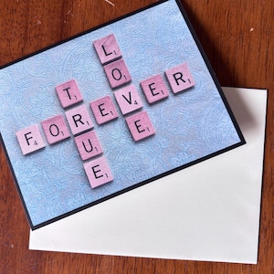 May include: A greeting card featuring the words "TRUE LOVE FOREVER" spelled out with pink Scrabble tiles on a blue patterned background. The card rests on a white envelope, set against a wooden surface.