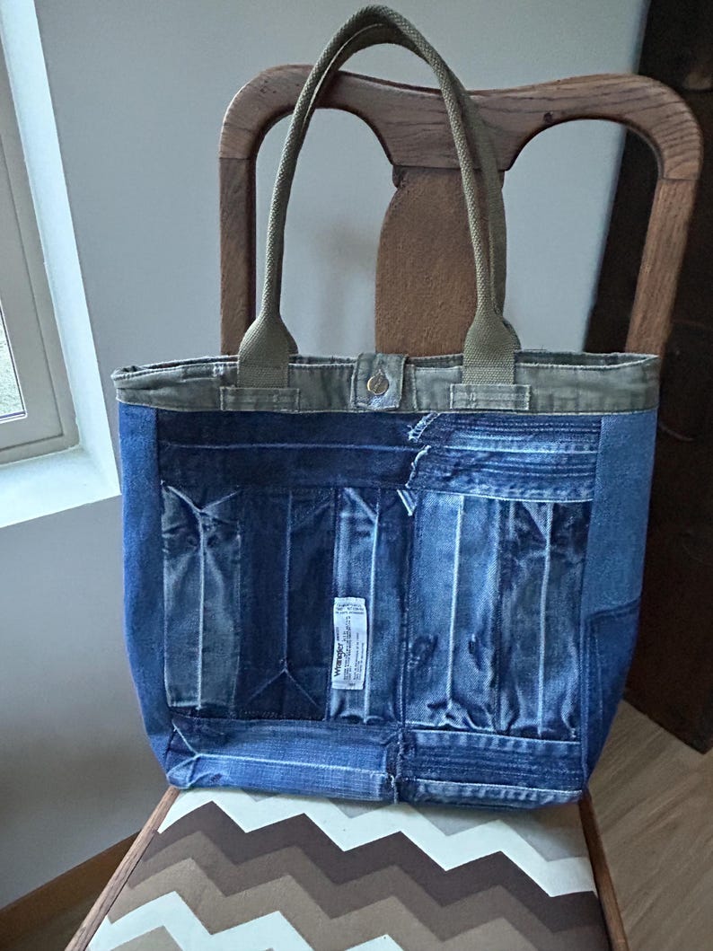 Puede incluir: Un bolso tote de mezclilla con detalles y asas de color verde oliva. El bolso est&aacute; hecho de mezclilla reciclada, con bolsillos y una etiqueta Wrangler. El bolso est&aacute; sobre una silla estampada.