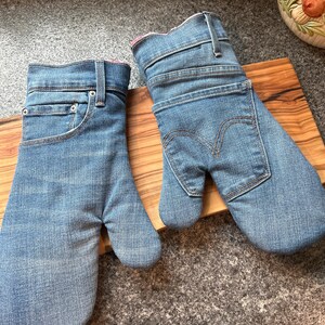 May include: A pair of oven mitts crafted from light blue denim fabric. One mitt features the front pocket and waistband details of jeans, while the other displays the back pocket with signature stitching. Both have a pink interior lining.