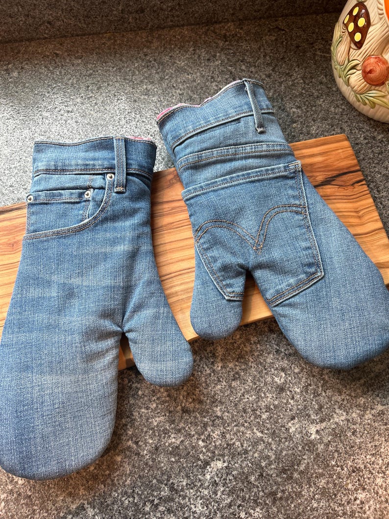 May include: A pair of oven mitts crafted from light blue denim fabric. One mitt features the front pocket and waistband details of jeans, while the other displays the back pocket with signature stitching. Both have a pink interior lining.