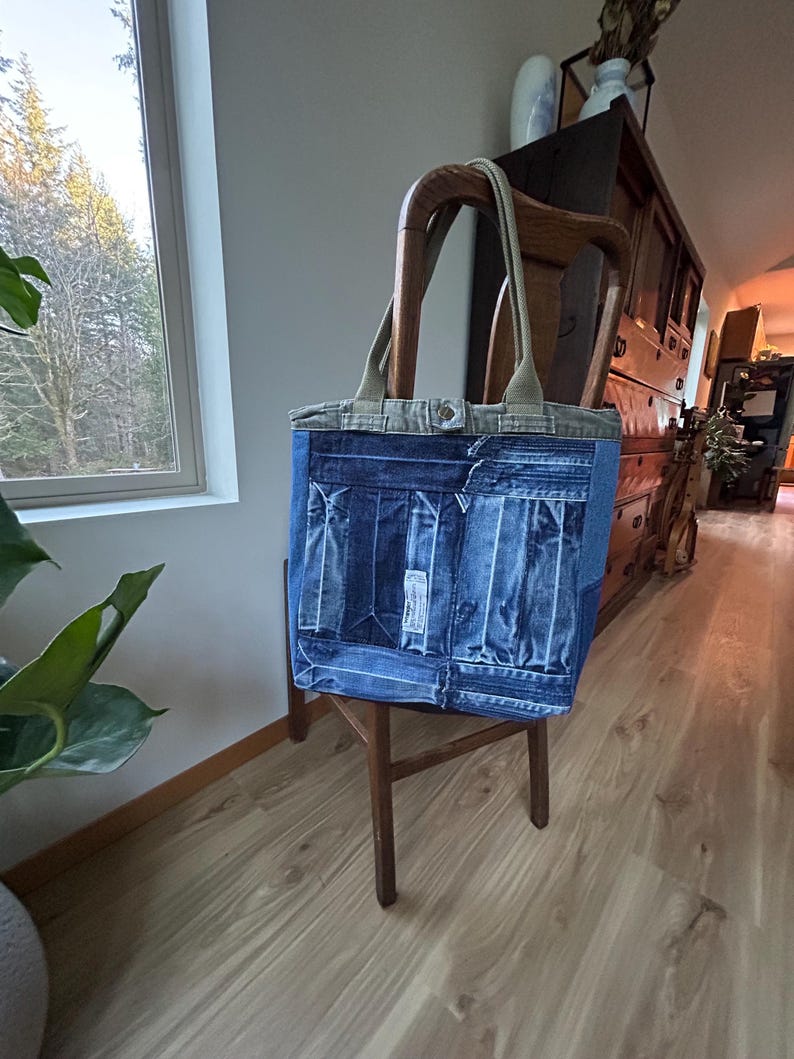 Puede incluir: Un bolso tote de mezclilla con un dise&ntilde;o de patchwork, con un cuerpo de mezclilla azul claro y una parte superior de mezclilla m&aacute;s clara con asas beige. La bolsa est&aacute; sobre una silla de madera en una habitaci&oacute;n bien iluminada.
