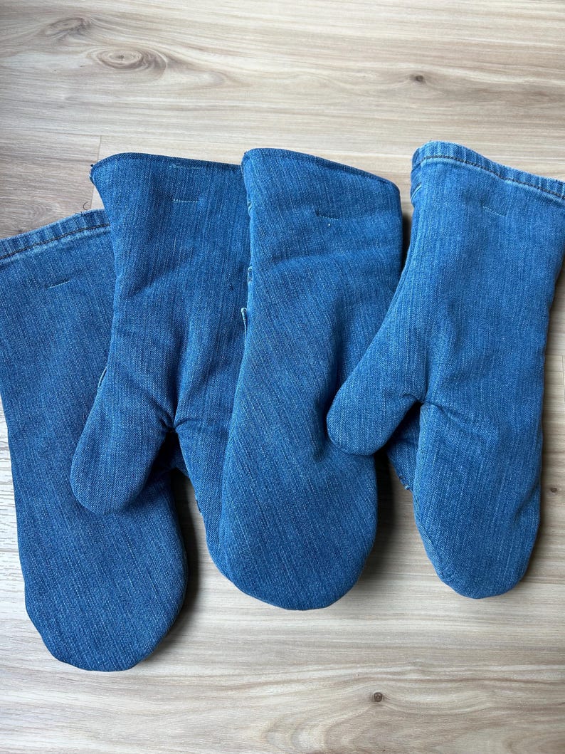 May include: Three oven mitts crafted from blue denim fabric. Each mitt features a standard thumb-separated design with a stitched hem at the opening. The denim material displays a consistent medium-blue wash with visible diagonal twill texture.