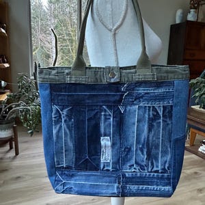 May include: A denim tote bag in shades of blue, with olive green handles and trim. The bag is constructed from recycled denim, featuring a patchwork design on the front. The handles are attached with metal hardware. The bag is displayed on a white stand.