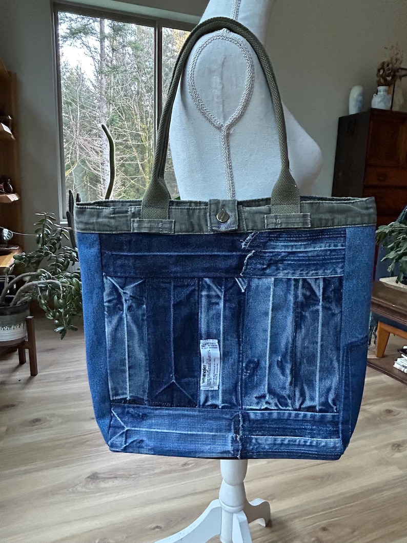Puede incluir: Un bolso tote de mezclilla azul con asas y ribetes verde oliva. El bolso est&aacute; hecho de mezclilla reciclada y presenta un dise&ntilde;o de patchwork en la parte delantera. Las asas est&aacute;n sujetas con herrajes met&aacute;licos. El bolso se exhibe sobre un soporte blanco.