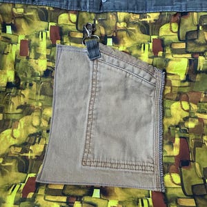 Puede incluir: Un bolso tote con un colorido estampado abstracto en tonos amarillos, verdes y marrones. Cuenta con un bolsillo beige y asas verde oliva. Un clip plateado est&aacute; sujeto al bolsillo.