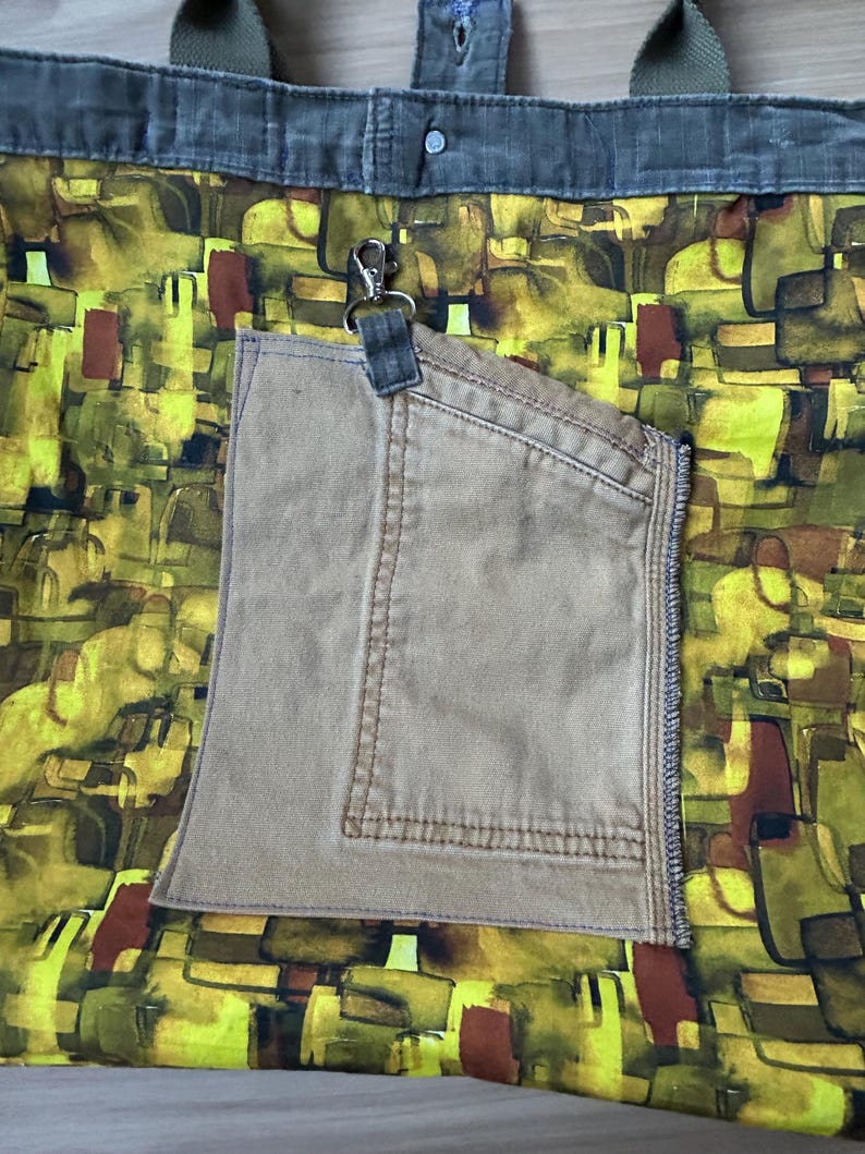 Puede incluir: Un bolso tote con un colorido estampado abstracto en tonos amarillos, verdes y marrones. Cuenta con un bolsillo beige y asas verde oliva. Un clip plateado est&aacute; sujeto al bolsillo.